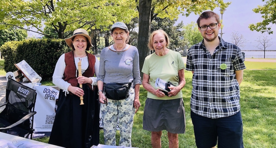 4 membres de la société se tiennent debout, une personnes avec une flute a bec dans les mains, dans un parc verdoyant, au soleil
