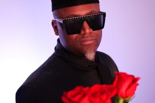Le chanteur pose avec des lunettes noires et un bouquet de roses rouges devant un fond violet